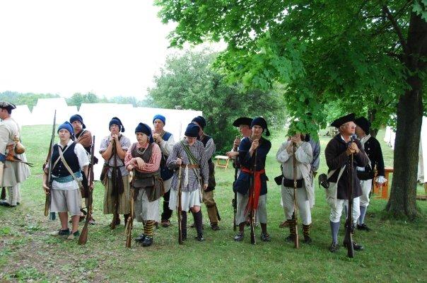 Militia and camp followers in period dress standing with muskets before white canvas tents