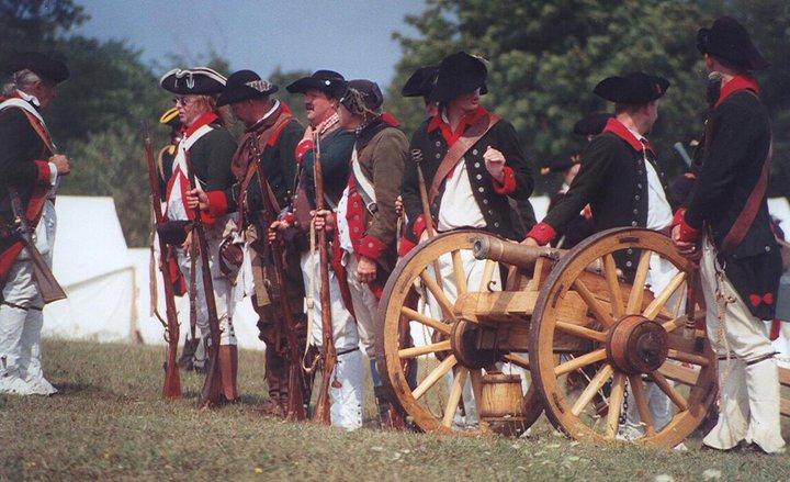 Re-enactors in green coats with red facings standing beside a small field cannon