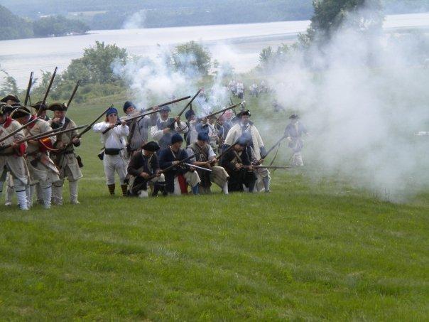 Two ranks of re-enactors firing muskets, white powder smoke drifting across the hillside