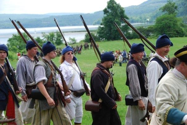 Militia line in 18th-century dress marching across a field with water and hills beyond